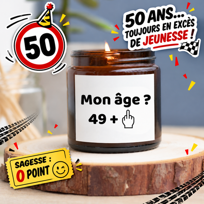 Bougie allumée en pot ambré avec le message Mon âge ? 49 + et un doigt d’honneur, entourée d’un panneau 50 et de confettis rouges et jaunes.