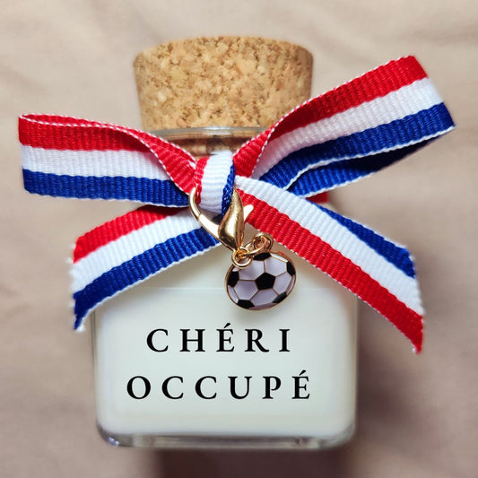 Bougie parfumée humoristique avec l'inscription "Chéri occupé" entourée de motifs de football, allumée dans un salon.