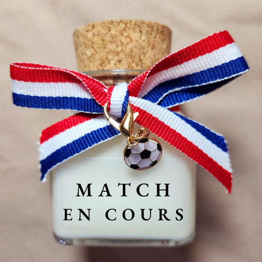 Bougie parfumée de supporter avec l'inscription Match en cours imprimée sur l'étiquette, allumée à côté d'une télé un soir de football.