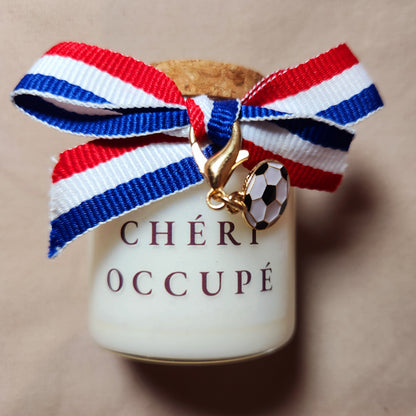 Bougie parfumée en verre avec l'inscription "Chéri Occupé" et des dessins de football, allumée sur une table basse en bois pendant une soirée match devant la télévision.