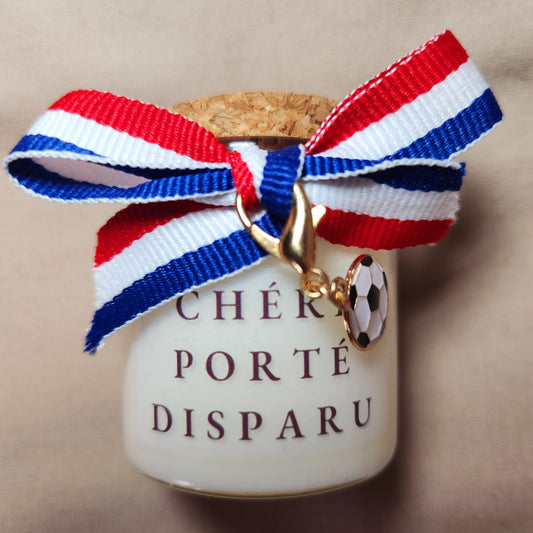 Bougie parfumée humoristique avec l'inscription "Chéri porté disparu" et des petits dessins de ballons de foot, allumée sur un meuble.