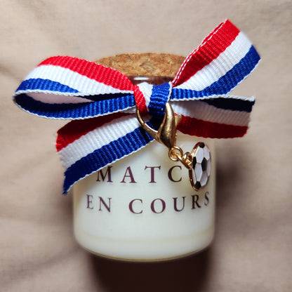 Bougie parfumée avec l'inscription "Match en cours" entourée de motifs de ballons de foot, allumée sur une table de salon.