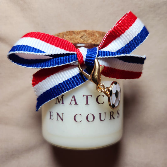 Bougie parfumée avec l'inscription "Match en cours" entourée de motifs de ballons de foot, allumée sur une table de salon.