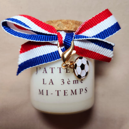 Bougie parfumée artisanale portant l'inscription coquine "J'attends la troisième mi-temps" avec des illustrations de football, allumée dans une ambiance de chambre ou de salon cosy.
