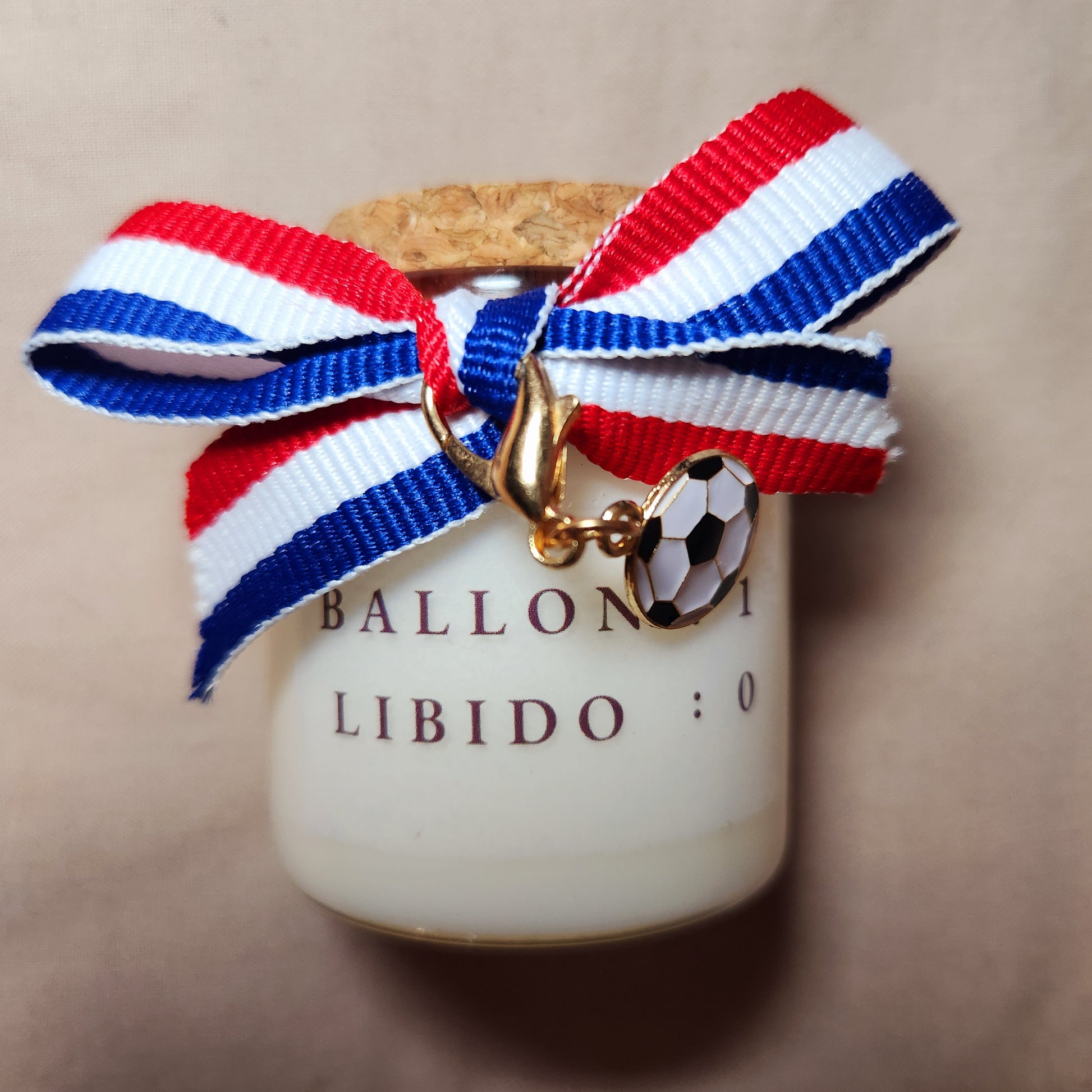 Bougie parfumée avec l'inscription humoristique "Ballon 1, Libido 0" écrite en noir, allumée sur un meuble avec un fond de salon tamisé.