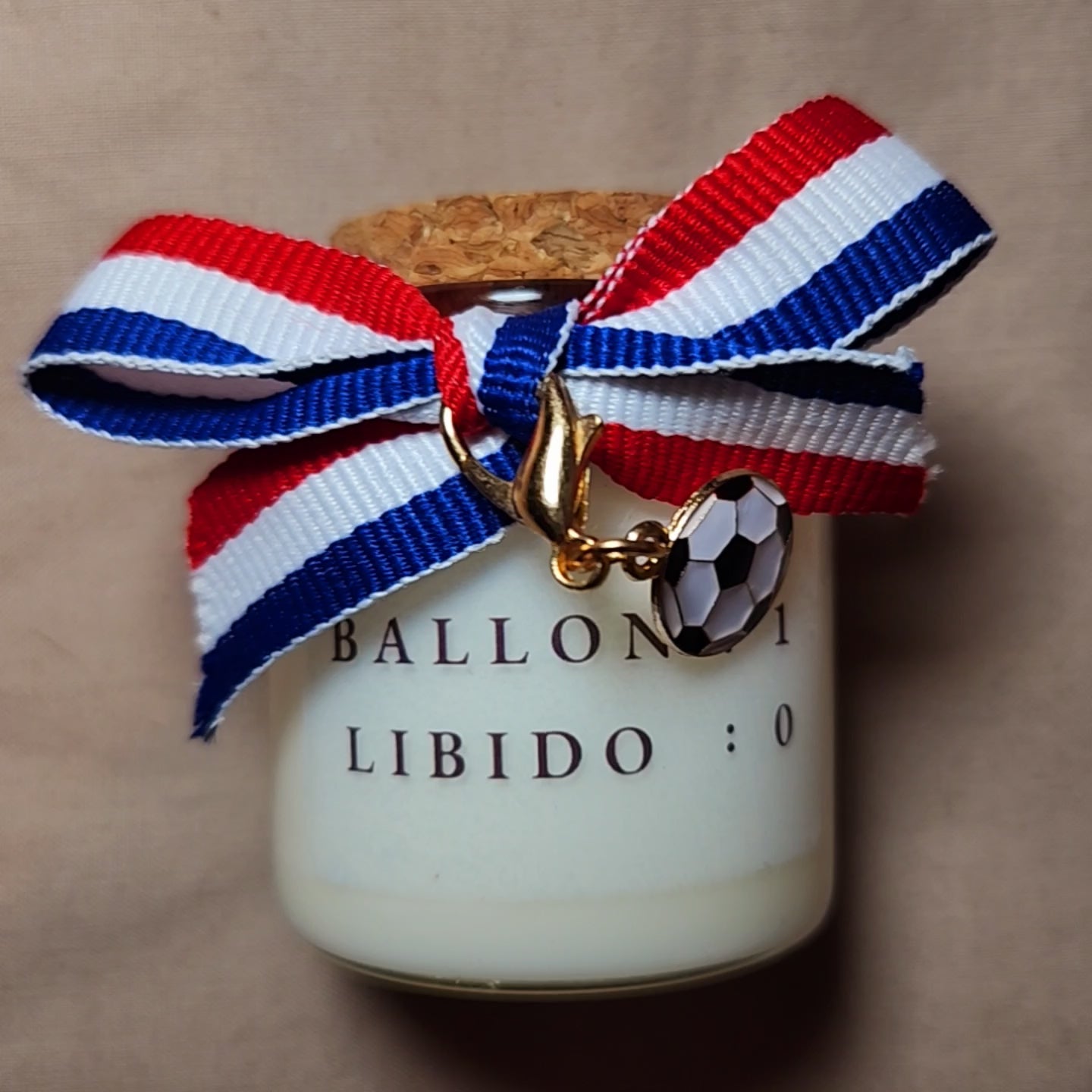 Bougie parfumée avec l'inscription humoristique "Ballon 1, Libido 0" écrite en noir, allumée sur un meuble avec un fond de salon tamisé. mamiecandle