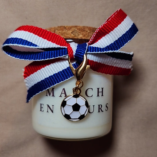 Bougie parfumée avec l'inscription "Match en cours" entourée de motifs de ballons de foot, allumée sur une table de salon mamiecandle