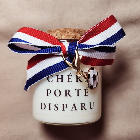 Bougie parfumée humoristique avec l'inscription "Chéri porté disparu" et des petits dessins de ballons de foot, allumée sur un meuble. mamiecandle